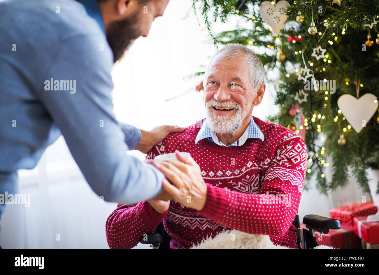 Un homme hipster méconnaissable en donnant une boisson dans une tasse à ses hauts père en fauteuil roulant à l'époque de Noël. Banque D'Images