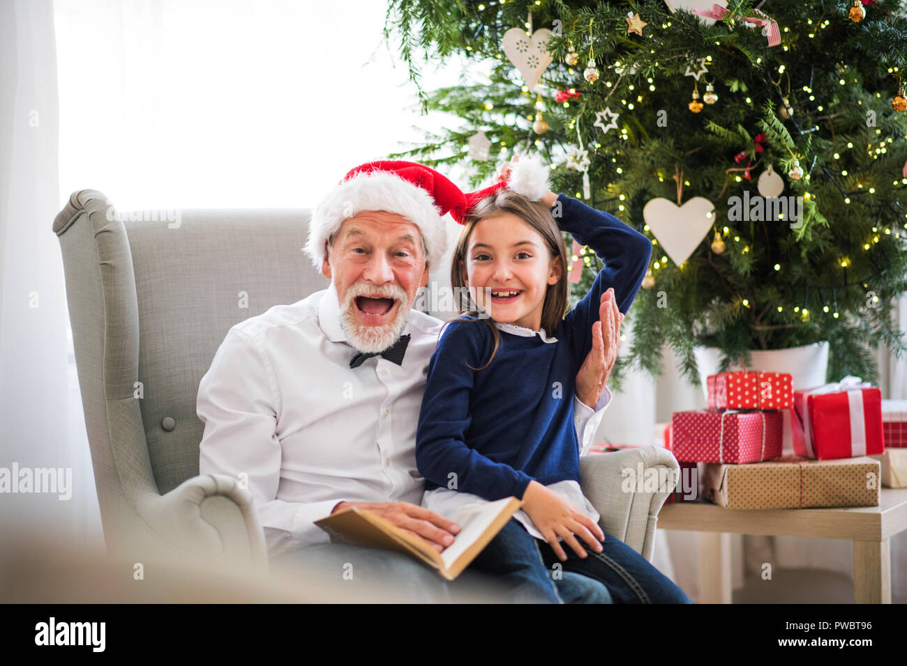 Une petite fille et de son grand-père avec Santa hat assis sur un fauteuil et de lire un livre à l'époque de Noël, plaisir. Banque D'Images