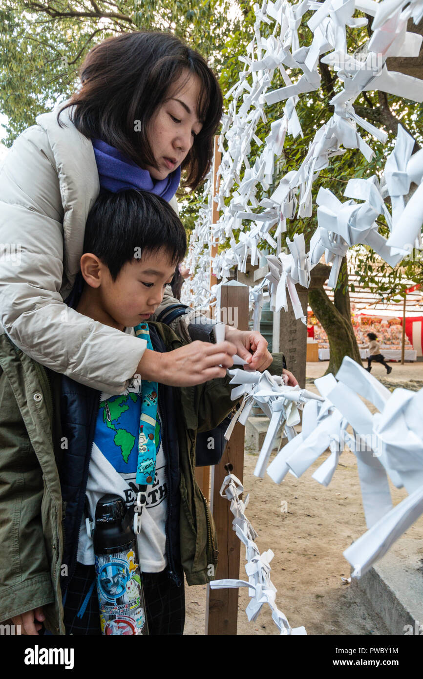 Nouvel An japonais, shogatsu. Jeune femme enfant aidant, garçon, 5-6 ans, de lier la mauvaise fortune de papier, Omikuji de cadre au sanctuaire de Nishinomiya. Banque D'Images