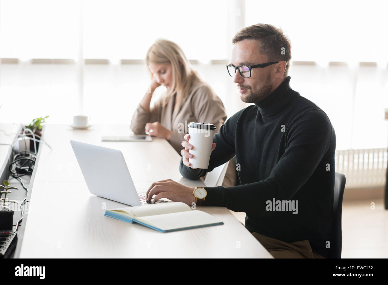 De graves pensive young bearded freelancer dans polo cou assis à table et de boire du café pendant l'utilisation d'ordinateur portable en espace de coworking Banque D'Images