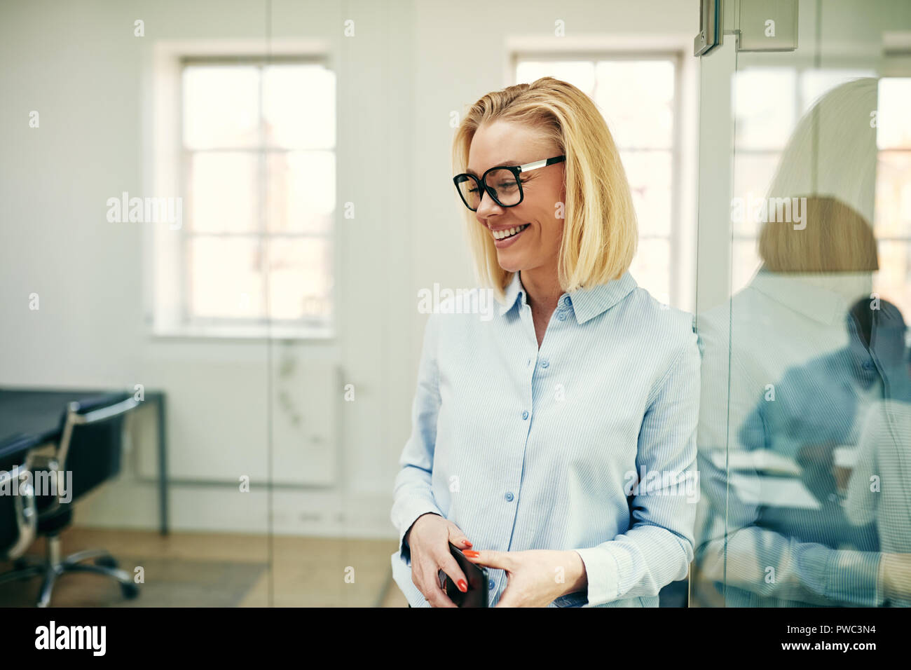 Confiant young businesswoman wearing glasses debout avec son téléphone cellulaire dans un bureau avec des collègues de travail à l'arrière-plan Banque D'Images
