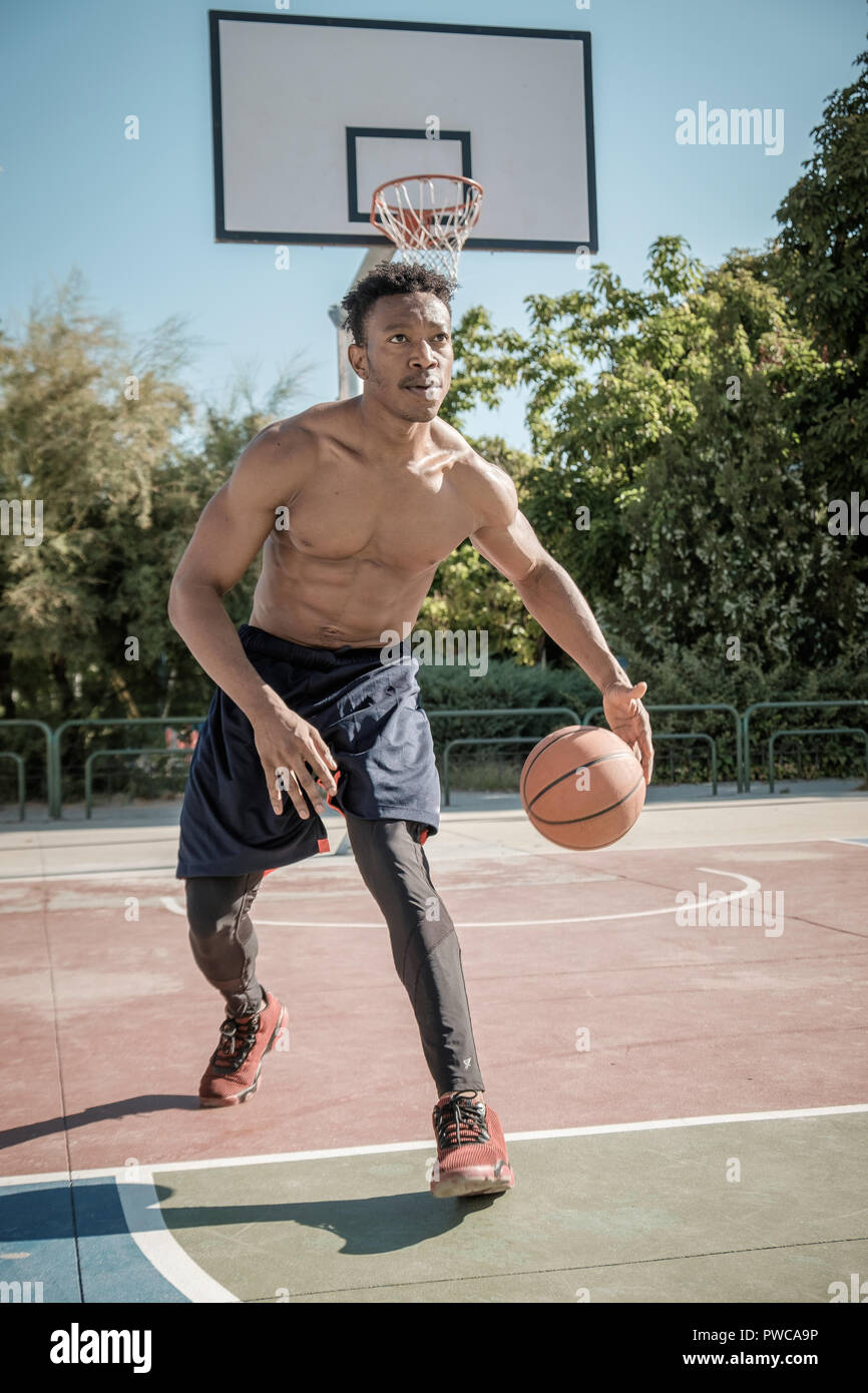 Un jeune homme afroamerican sans tshirt est jouer au basket-ball dans un  parc de Madrid au cours de l'été à midi. Il est de rebondir la balle sous  le panier Photo Stock -