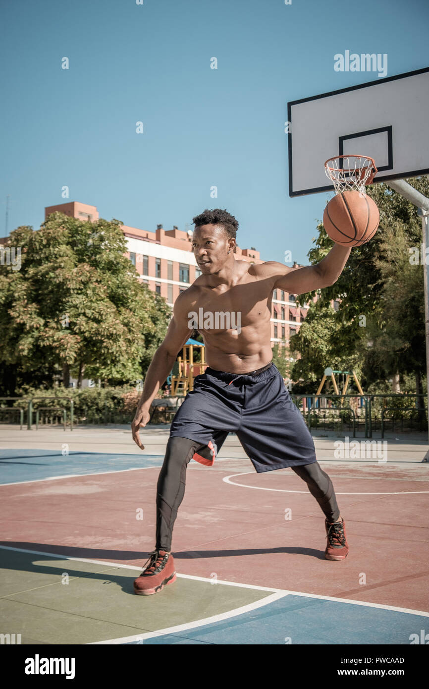 Un jeune homme afroamerican sans tshirt est jouer au basket-ball dans un parc de Madrid au cours de l'été à midi. Il est de rebondir la balle sous le panier. Banque D'Images