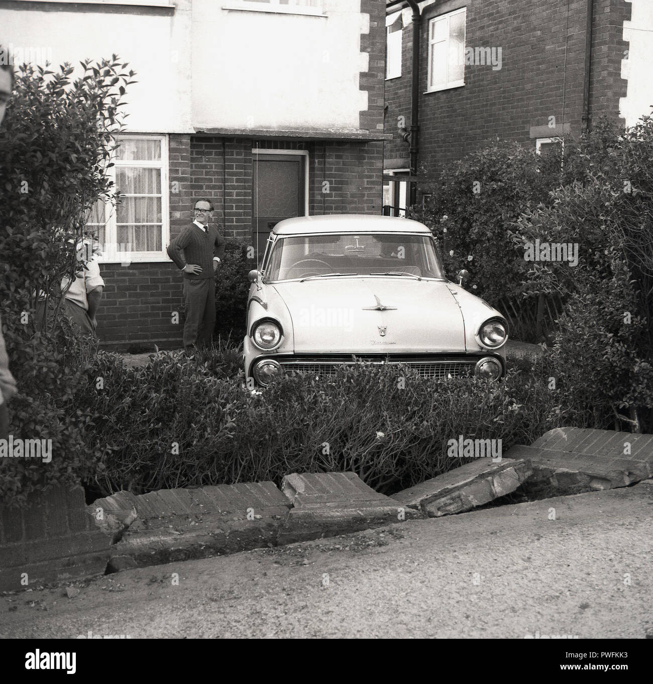 1967, comment est-ce arrivé là ?, un homme pensif devant sa porte de sa chambre se trouve une direction à droite, de style transatlantique britannique motor car assis dans son petit jardin à l'avant, ayant été (renversé) à travers un mur de briques, Hardwick, Buckinghamshire, Angleterre, Royaume-Uni. Banque D'Images