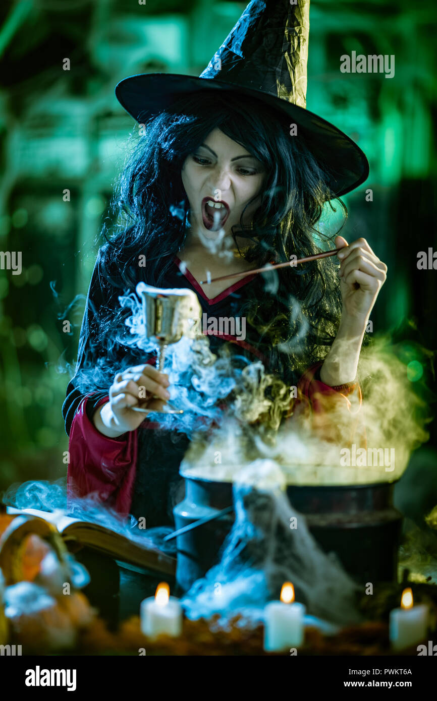 Jeune sorcière avec visage sérieusement en environs et fumé fond vert est titulaire d'un gobelet avec potion magique. Banque D'Images