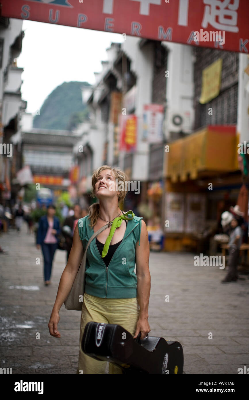 Young Woman smiling en marchant le long d'une rue portant un étui à guitare. Banque D'Images