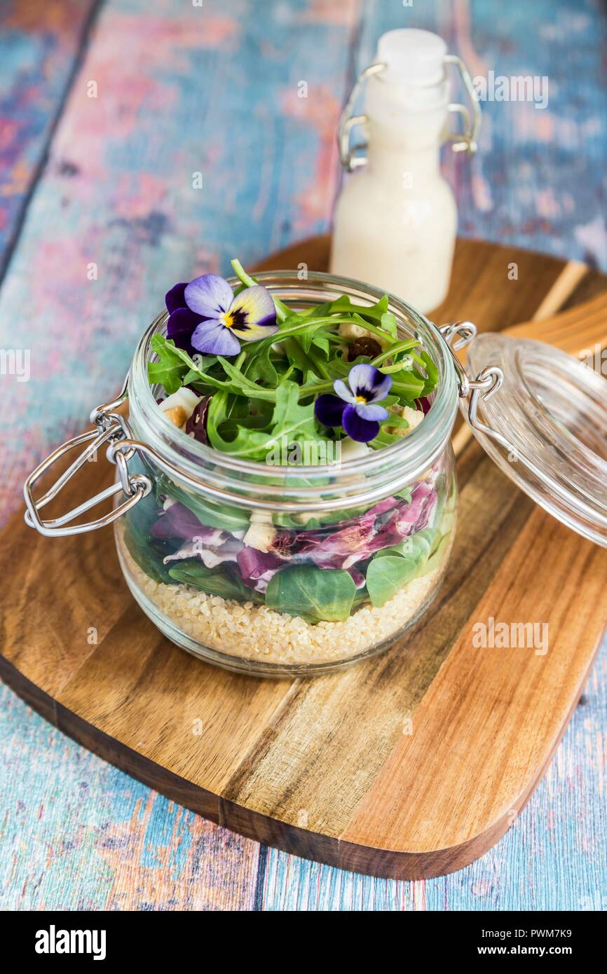 Une Salade de quinoa aux agneaux avec laitue, chicorée, roquette, croûtons, fromage de chèvre et violettes cornu dans un bocal en verre sur une planche en bois, avec la vinaigrette dans un Banque D'Images