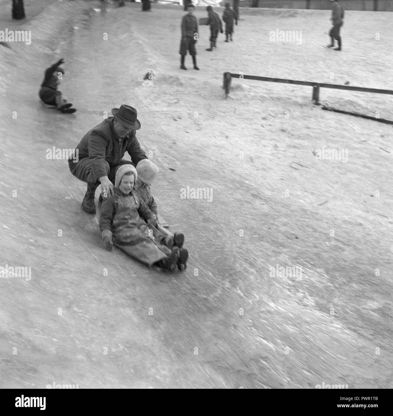 L'hiver dans les années 1950. Des enfants jouent dans un parc à Stockholm. Le glisser sur la colline glacée ensemble et avoir du plaisir. Même un homme adulte est dans le jeu. Suède 1951. Ref 1609 Banque D'Images