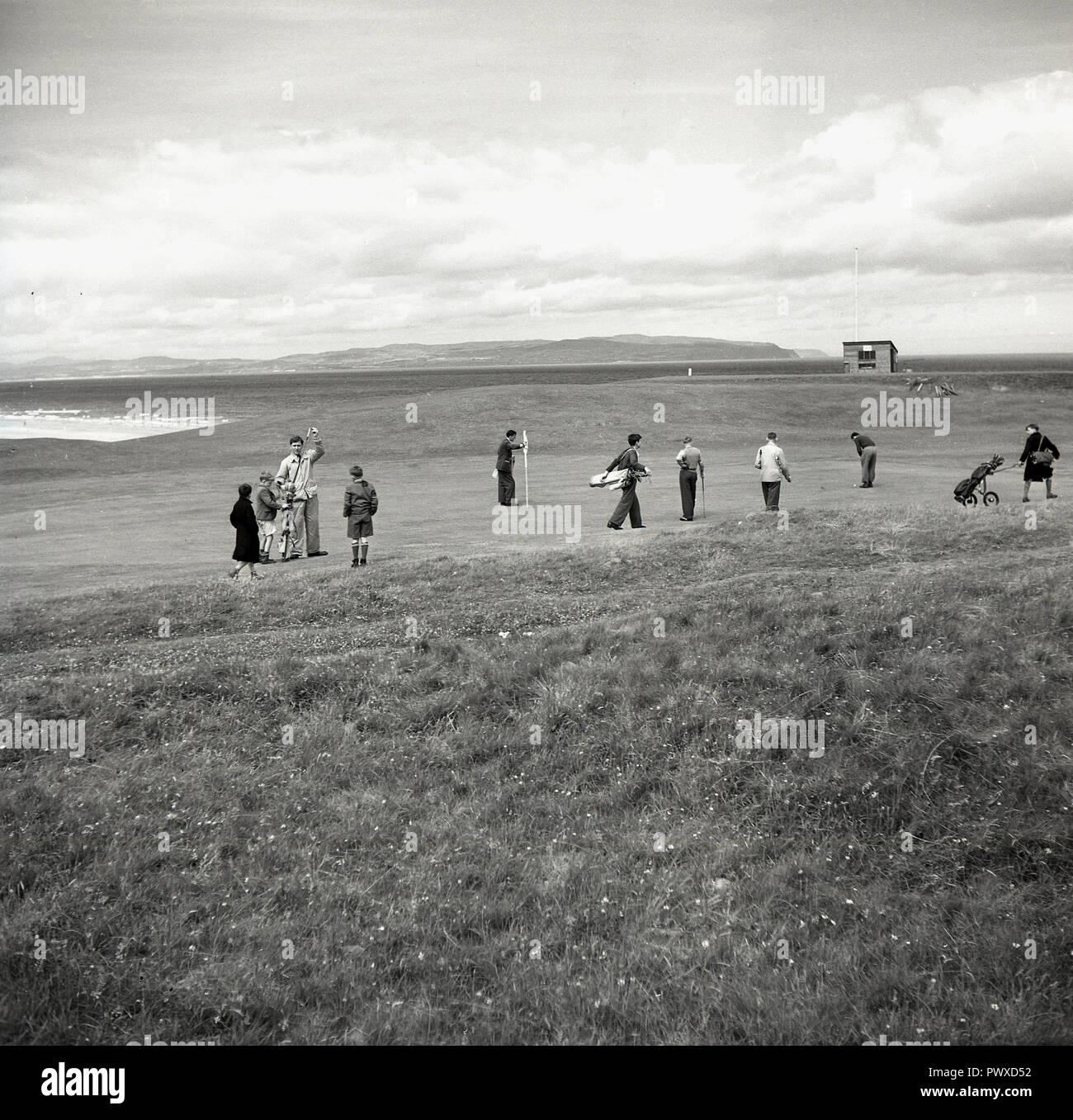 Années 1950, historiques, golf, golf, golfeurs masculins jouant sur un parcours côtier sur le putting green, avec leurs petits caddies présents en Irlande du Nord, Royaume-Uni. Banque D'Images