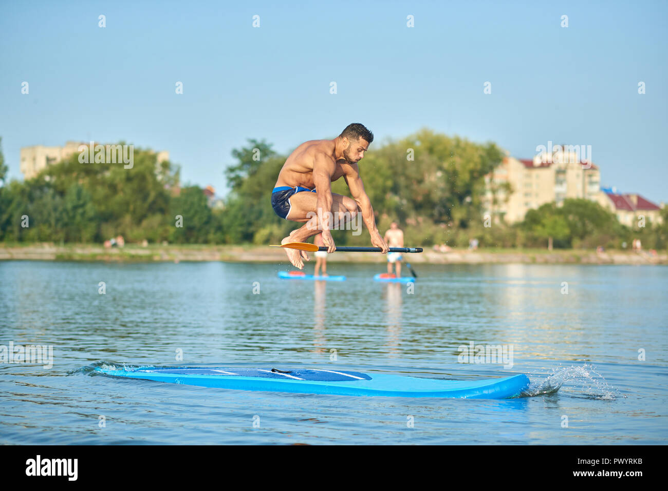 Handsome man training musculaire on paddle board, sautant au-dessus de lui avec longue rame dans la main au lac de la ville. Jeune garçon fit porter des shorts de sport, l'exercice sur l'eau. Concept de sport et mode de vie sain. Banque D'Images