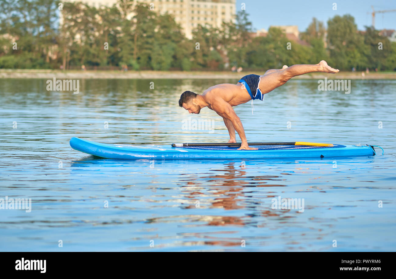 Muscular man practicing yoga, faisant équilibre de poids de corps, la natation on paddle board au milieu du lac de la ville, au cours de journée ensoleillée. Sportsman avec figure parfaite, vêtu d'un short bleu, la formation à l'extérieur. Banque D'Images