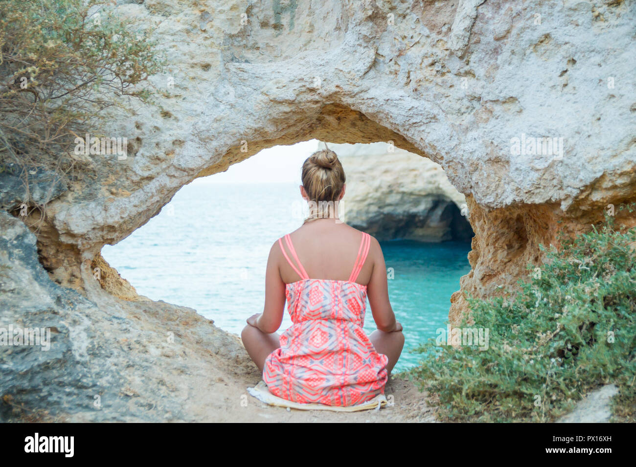 Jeune femme pratiquant la méditation pendant le coucher du soleil sous une arche de pierre calcaire naturelle par la formation d'un lagon turquoise sur l'Algarves, Portugal. Banque D'Images