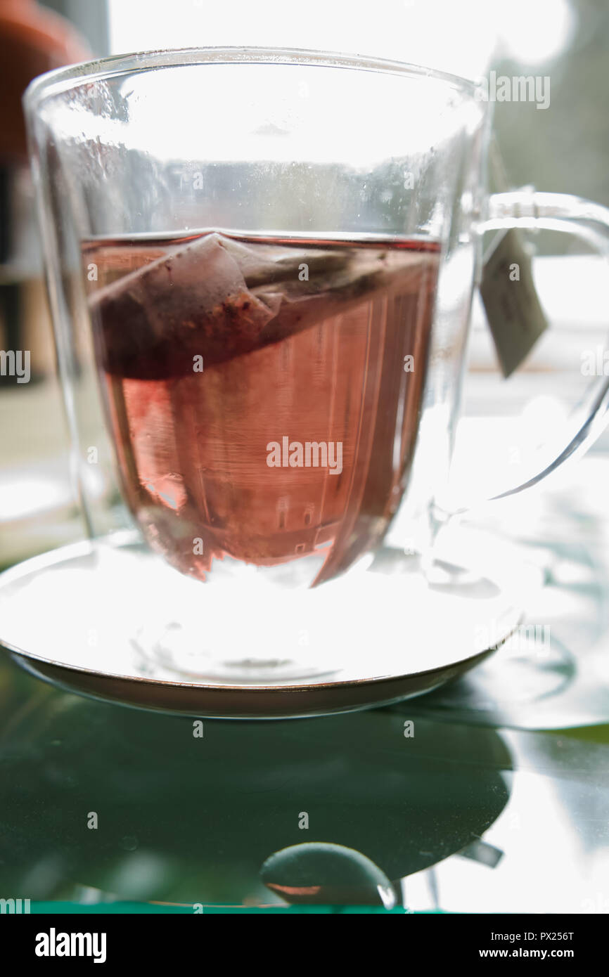 Un sachet de thé noir donne à la tasse en verre une couleur rougeâtre. Banque D'Images