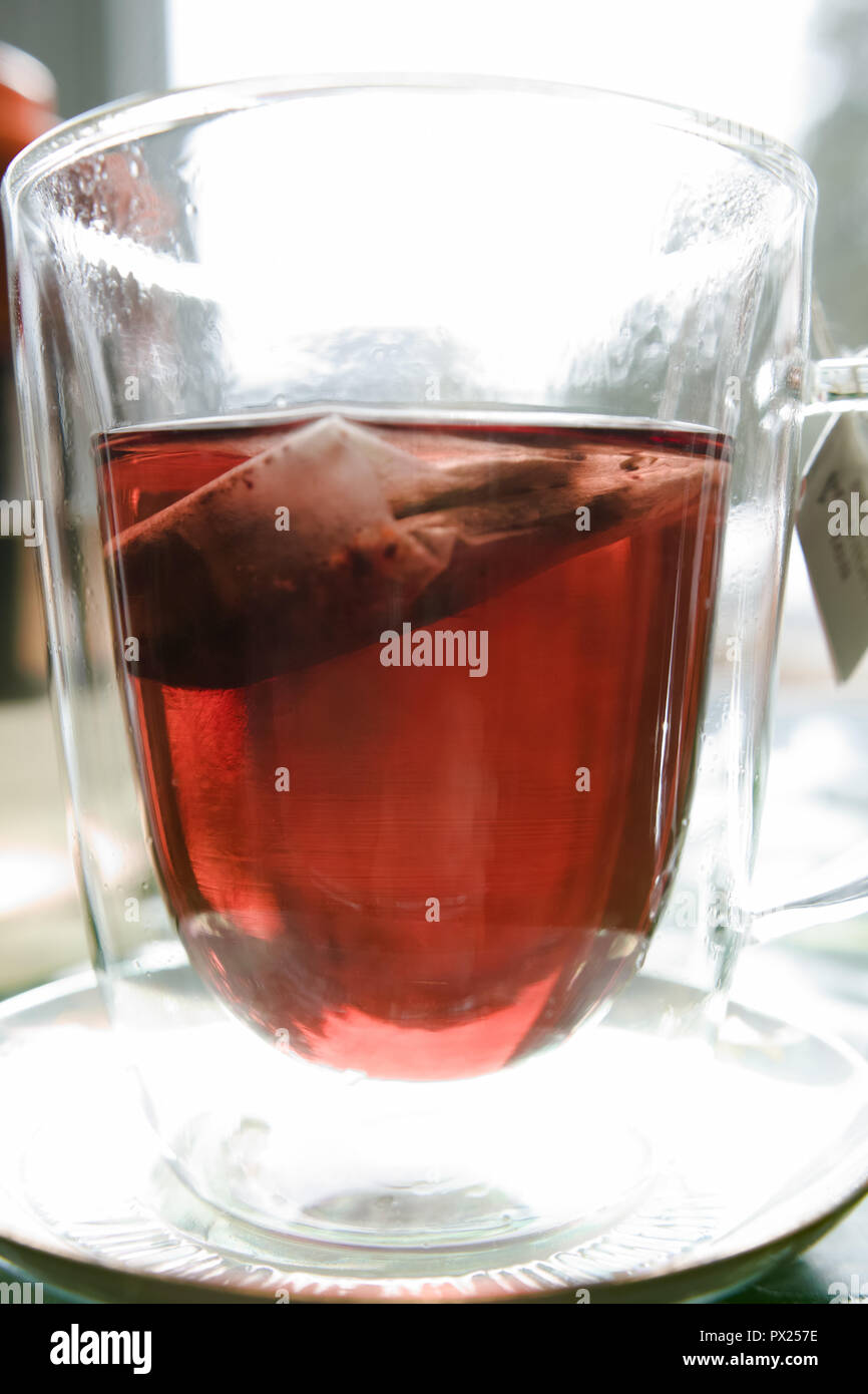 Un sachet de thé noir donne à la tasse en verre une couleur rougeâtre. Banque D'Images