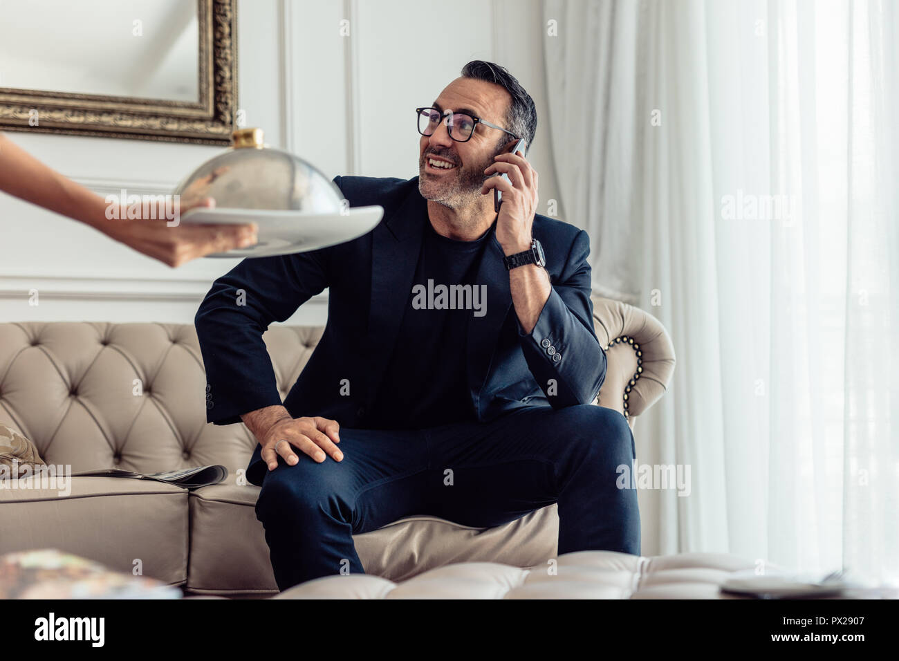 Mature businessman sitting on sofa talking on mobile phone in hotel room avec un serveur servant de la nourriture. Service en chambre garçon servant de la nourriture à l'homme d'affaires Banque D'Images