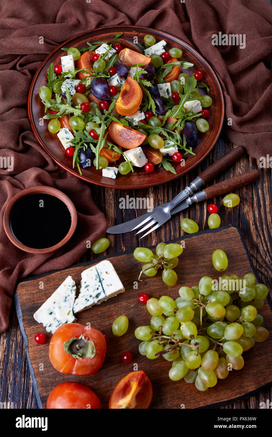 Salade d'automne délicieux avec du chocolat kaki, fromage bleu, raisin vert, tranches de prune et la roquette sur une plaque de faïence avec des ingrédients sur un cutti Banque D'Images