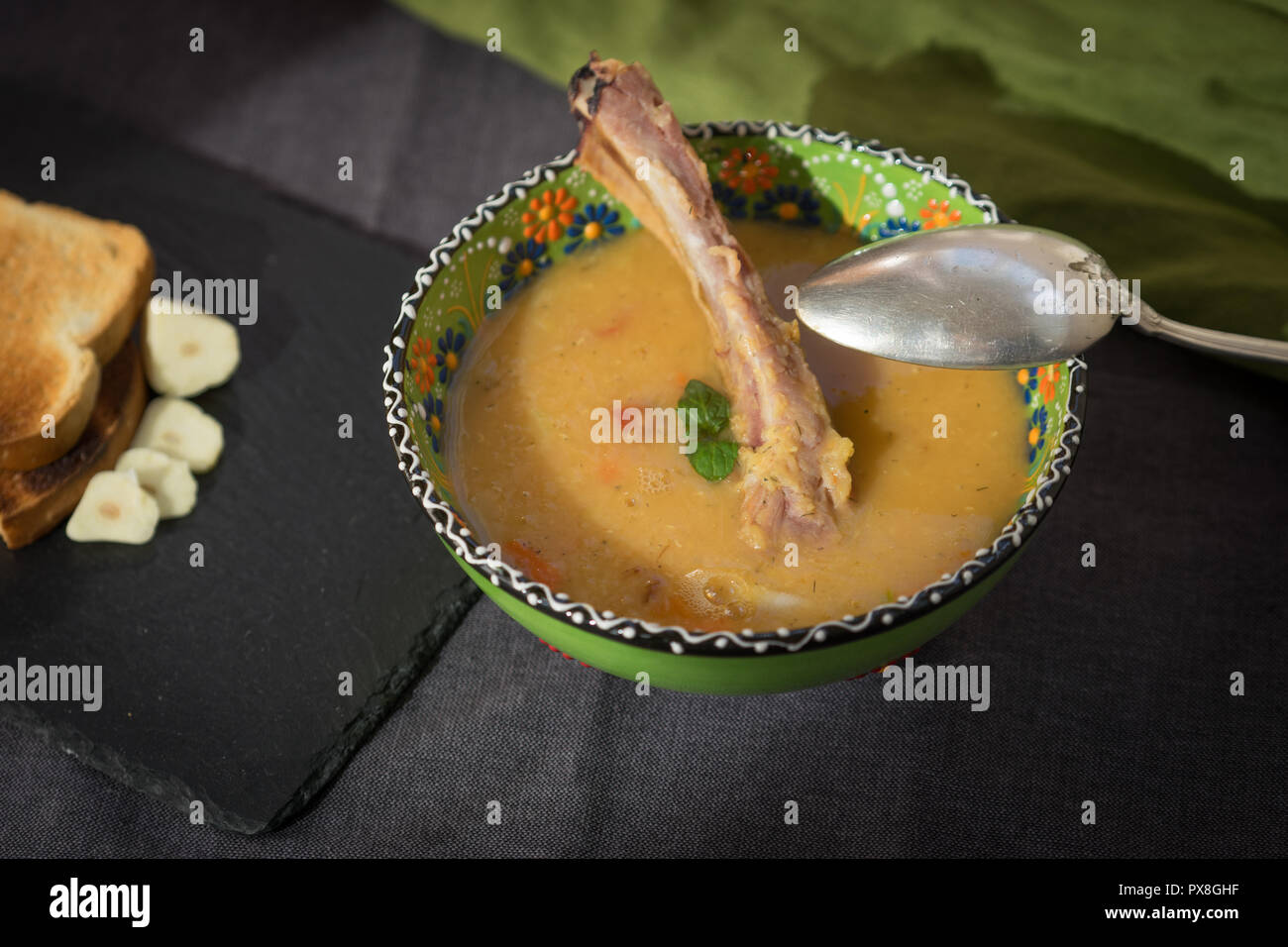 Soupe aux lentilles rouges avec côtes levées fumées dans un bol vert clair avec l'ornement floral. Fond tissu gris foncé. Banque D'Images