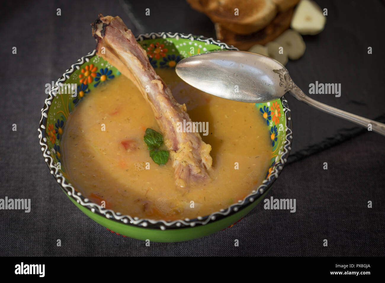 Soupe aux lentilles rouges avec côtes levées fumées dans un bol vert clair avec l'ornement floral. Fond tissu gris foncé. Banque D'Images