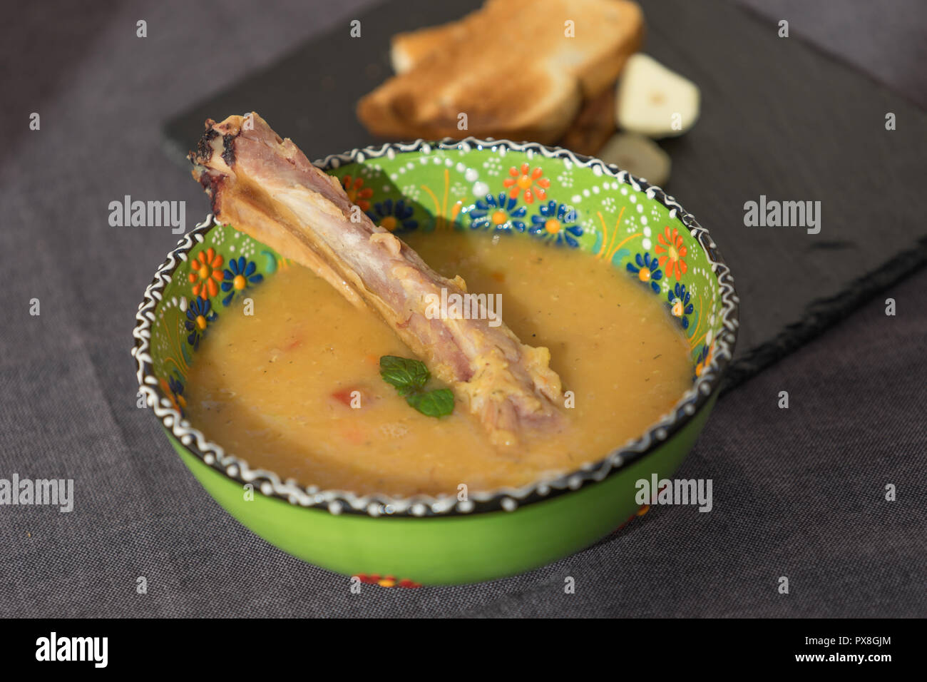 Soupe aux lentilles rouges avec côtes levées fumées dans un bol vert clair avec l'ornement floral. Fond tissu gris foncé. Banque D'Images