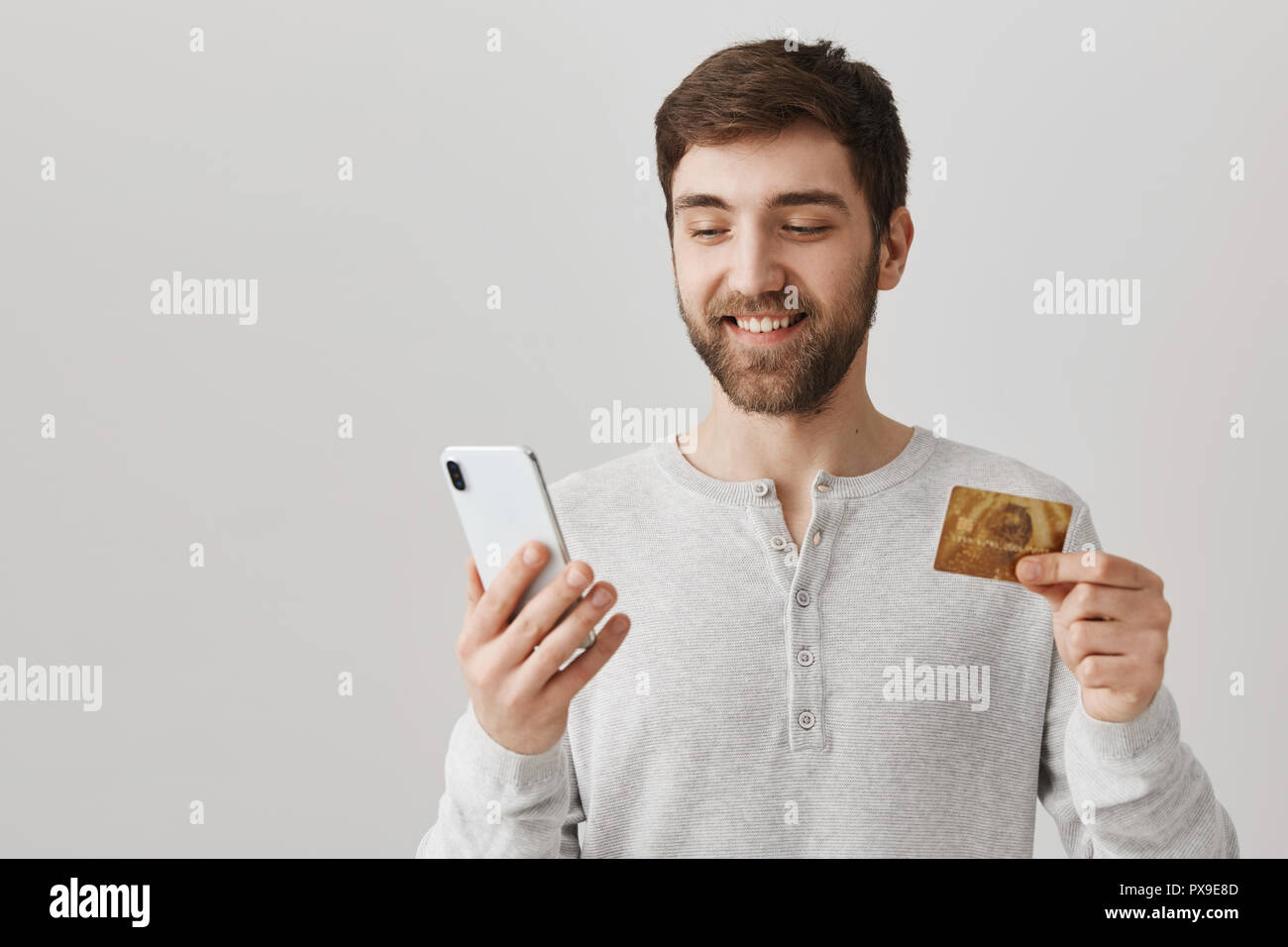 Heureux l'homme à barbe, holding smartphone et une carte de crédit, à la recherche à l'écran comme s'il n'est jusqu'à acheté quelque chose, debout sur fond gris. Guy veut impressionner copine et dress Banque D'Images