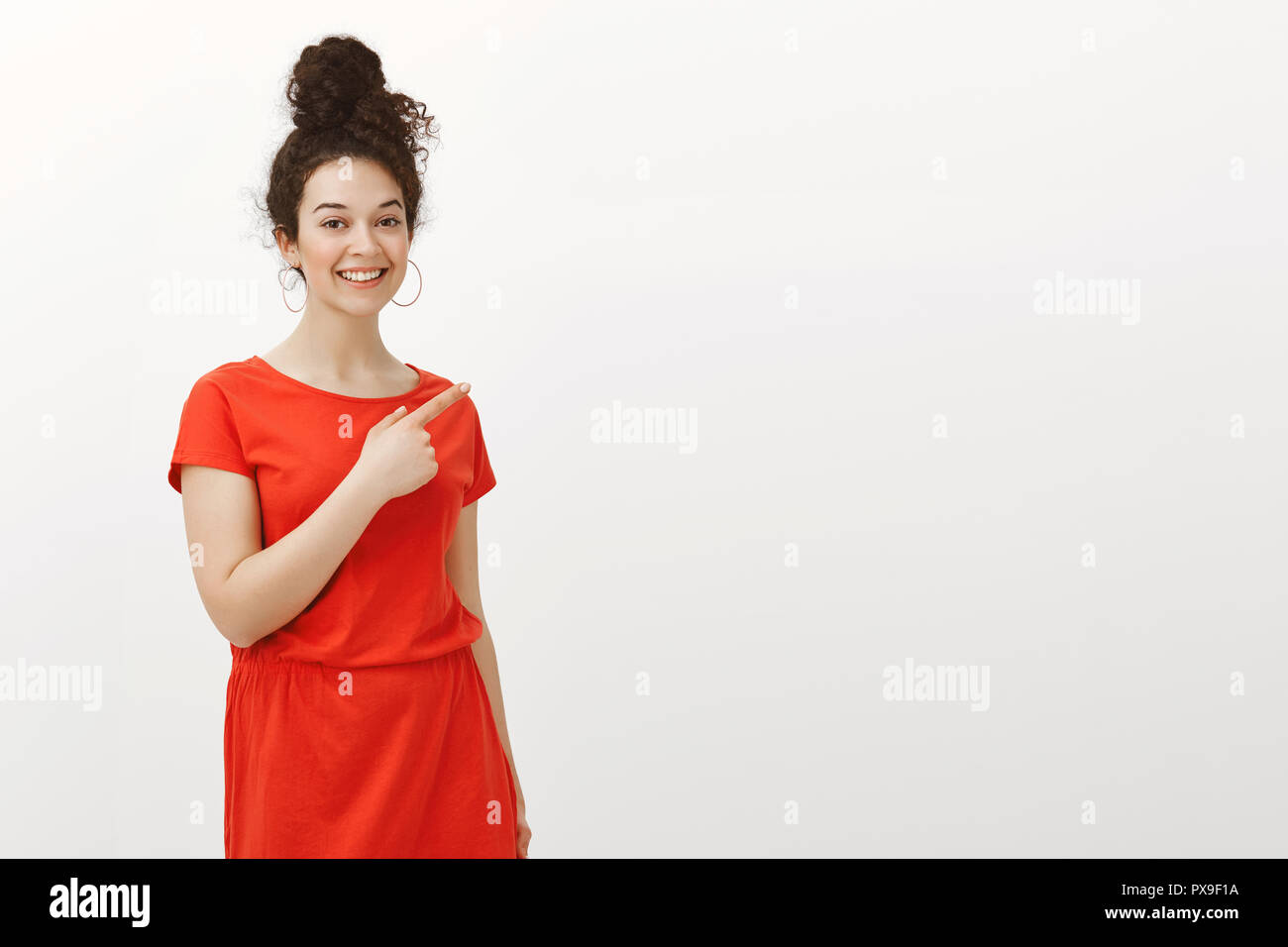 Portrait de plaisir jolie femme aux cheveux bouclés en belle robe, pointant vers le coin supérieur droit en souriant largement, l'expression de l'attitude sympathique et heureux, montrant sur fond gris Banque D'Images