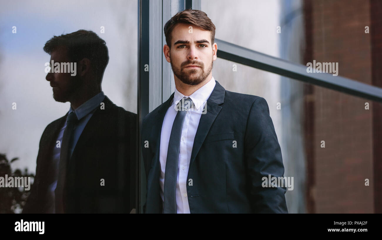 Portrait of handsome young man par bâtiment de bureaux et à regarder l'appareil photo. L'homme en costume d'affaires looking at camera while standing outsid Banque D'Images