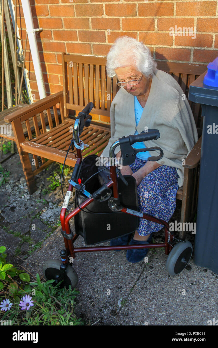 Une femme âgée assise sur un banc à l'extérieur de la marche à l'aide d'un chariot à roues, 3 poussette, 3 roues aide à la marche ou 3 roues walker, England, UK Banque D'Images