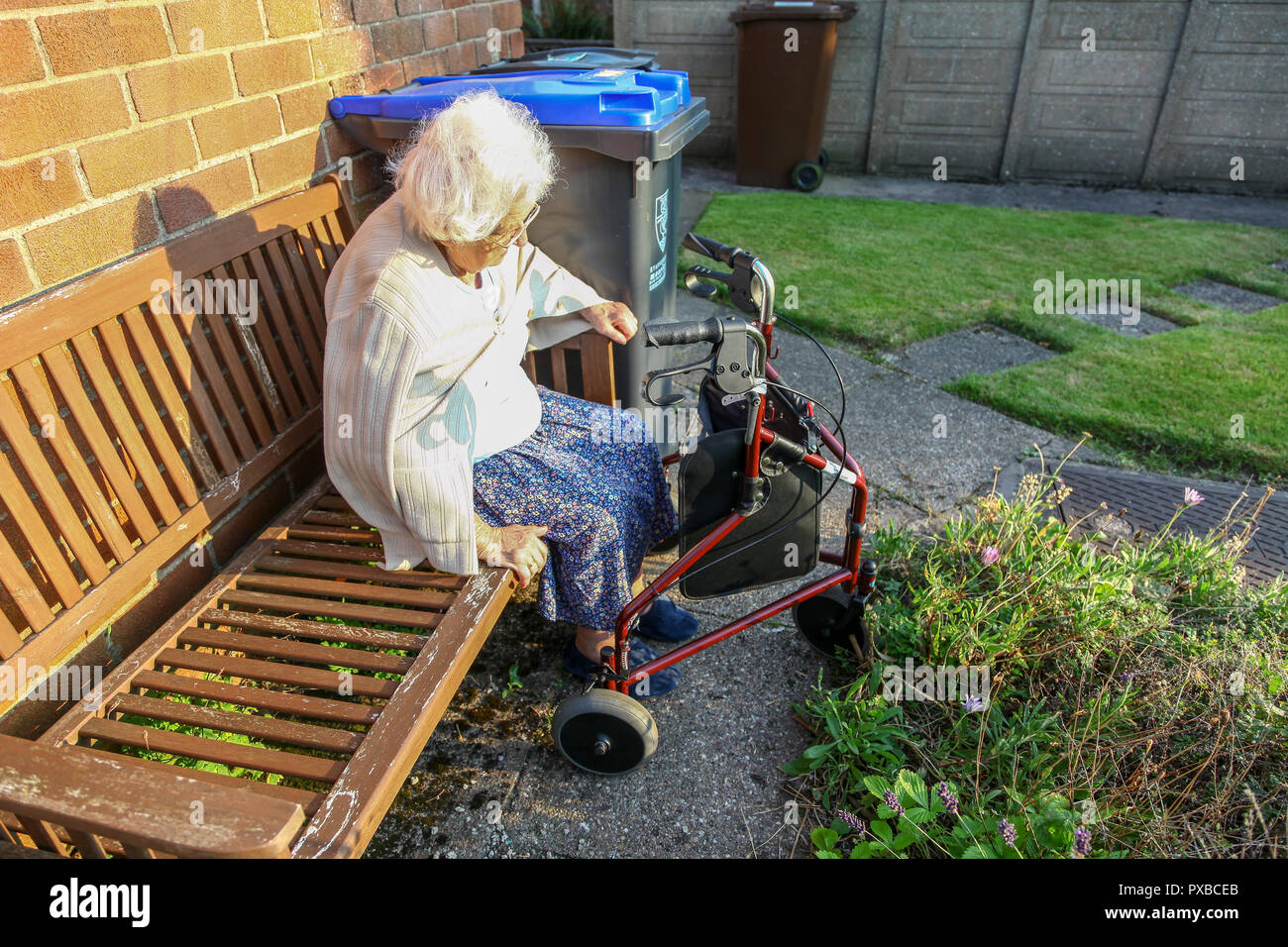 Une femme âgée assise sur un banc à l'extérieur de la marche à l'aide d'un chariot à roues, 3 poussette, 3 roues aide à la marche ou 3 roues walker, England, UK Banque D'Images