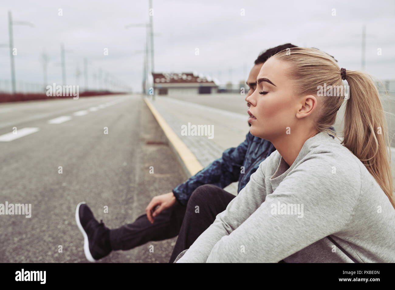 Jeune couple divers dans les vêtements de sport assis sur le bord d'une route ensemble en faisant une pause à partir d'une exécution sur un jour nuageux Banque D'Images