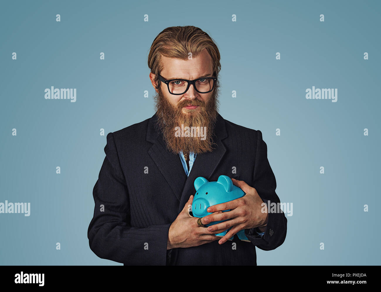 Jeune homme d'hipster barbu avare cupide man holding piggy bank isolé sur fond mur studio bleu. Face à l'émotion humaine expression négative Banque D'Images