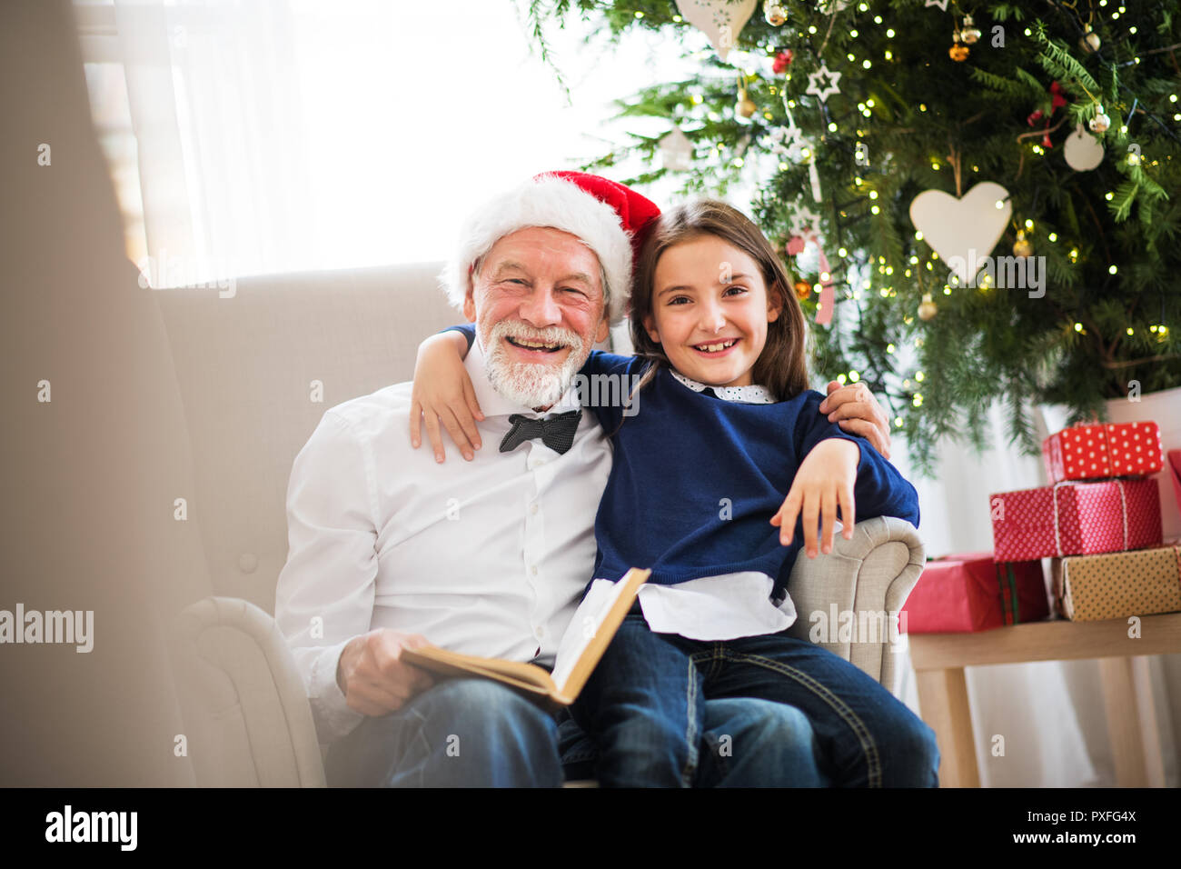 Une petite fille et de son grand-père avec Santa hat assis sur un fauteuil et de lire un livre à l'époque de Noël. Banque D'Images