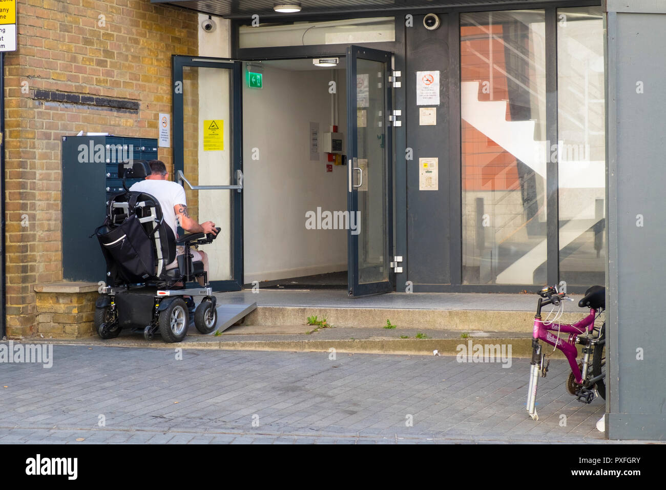 Homme dans un fauteuil roulant motorisé qui monte une rampe métallique dans un bâtiment, uk Banque D'Images