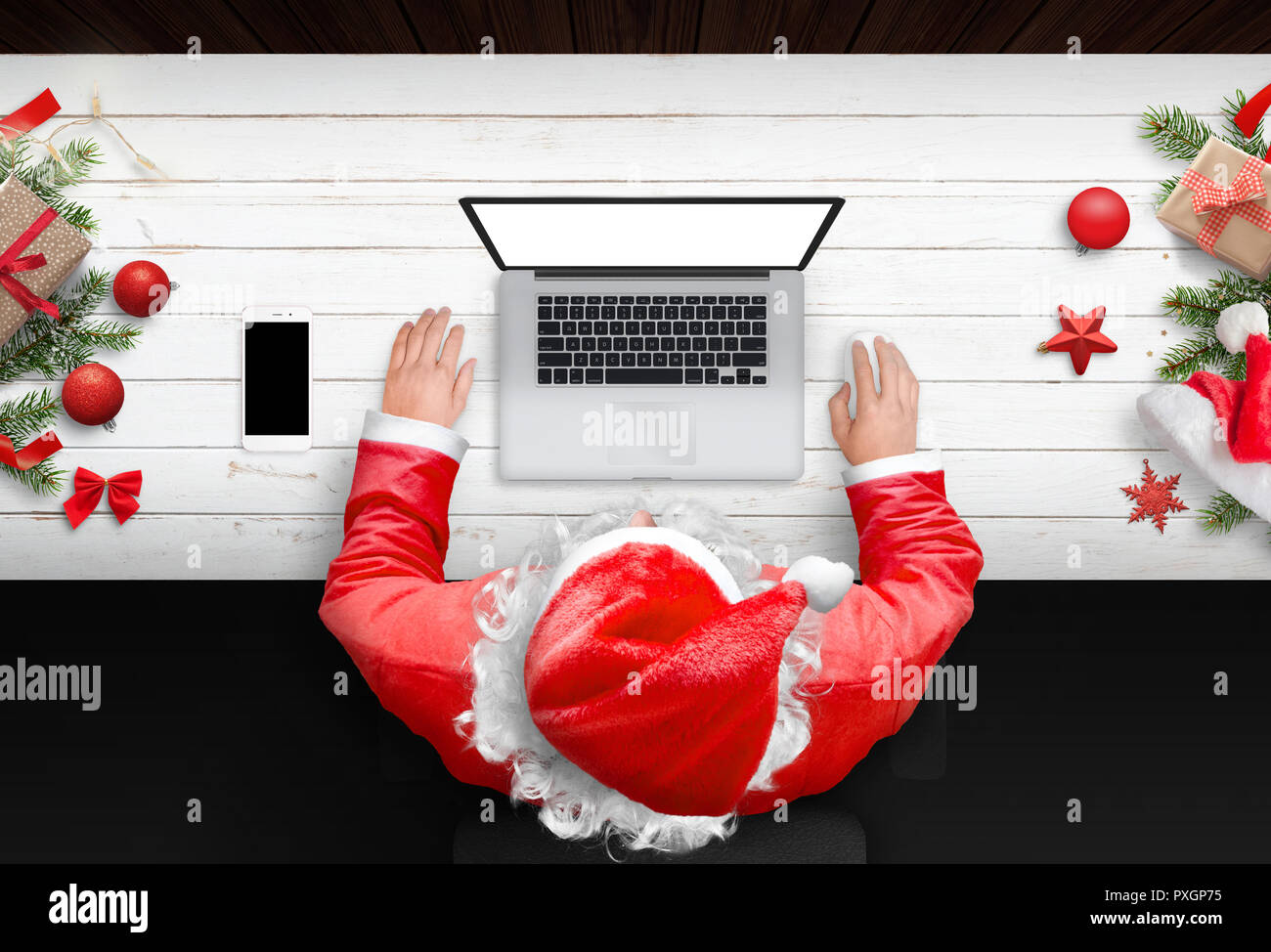 Santa Claus travailler sur ordinateur portable avec écran isolé pour une maquette. Le temps de Noël. Bureau en bois avec des décorations de Noël. Téléphone portable à côté. Haut Banque D'Images