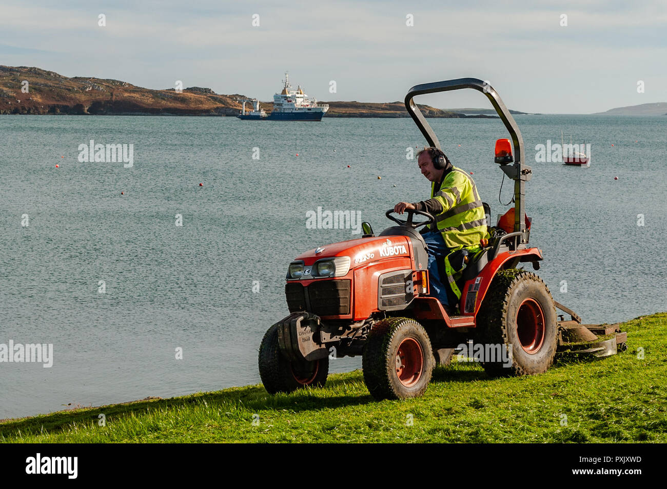Schull, West Cork, Irlande. 23 Oct, 2018. Un employé de Cork County Council coupe l'herbe sur Schull waterfront comme navire de pose de bouées 'Granuaile' se trouve à l'ancre au large de Schull. Sunshine a été l'ordre du jour aujourd'hui avec des hauts de 13 à 15° Celsius à West Cork. Credit : Andy Gibson/Alamy Live News. Banque D'Images