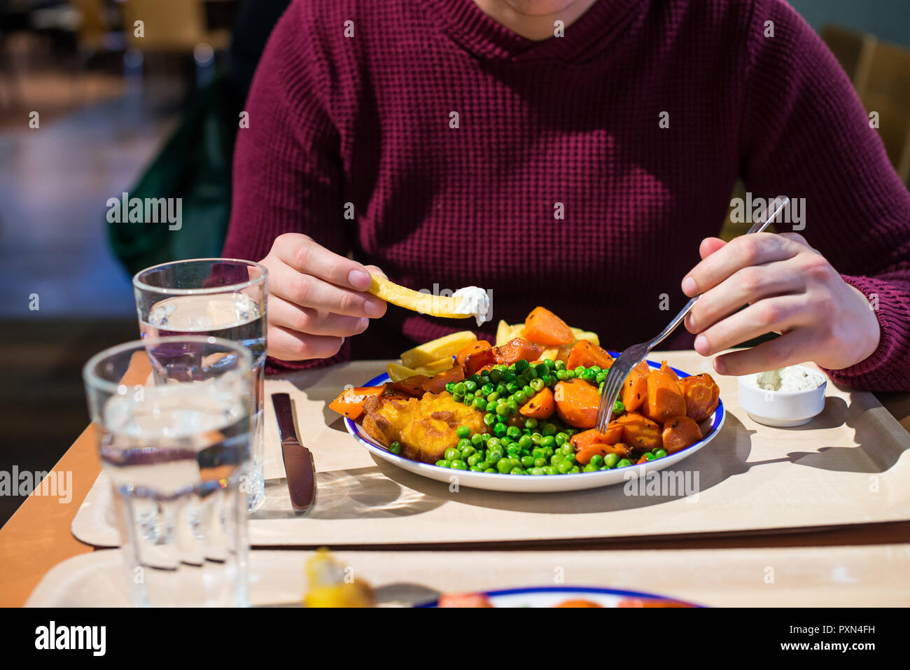 Jeune homme tenant couverts et manger un plat traditionnel anglais - fish and chips avec petits pois, carottes cuites et de tartre sause. Restauration rapide, café affaires Banque D'Images