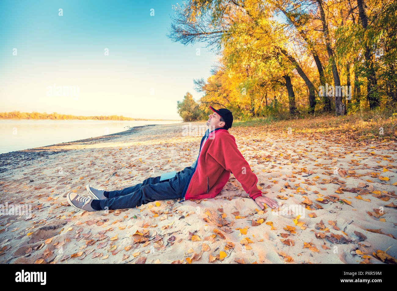 Un homme heureux avec les yeux fermé assis sur la rive du fleuve sur le sable avec des feuilles tombées en automne. L'homme en bonne météo ensoleillée Banque D'Images