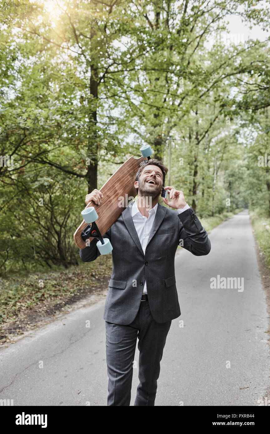 Happy businessman with skateboard parler sur smartphone on rural road Banque D'Images