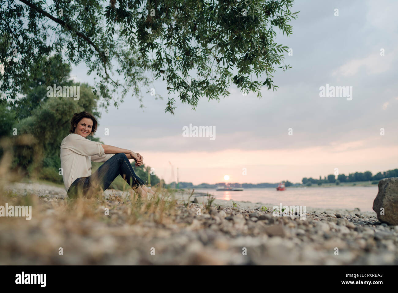 Femme assise au bord de la rivière, à regarder le coucher du soleil Banque D'Images