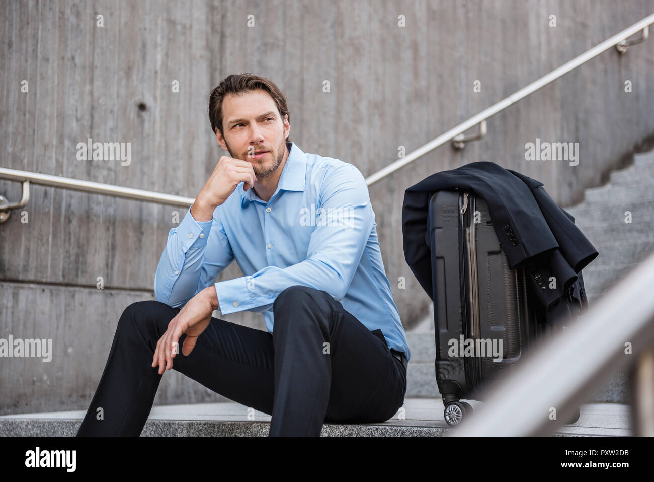 Businessman with rolling suitcase sitting on stairs Banque D'Images