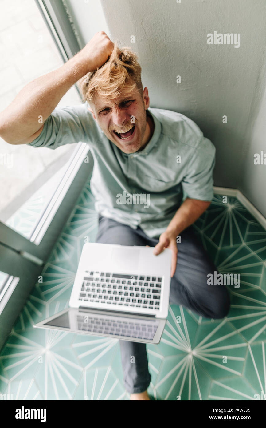 Jeune homme assis sur le sol, holding laptop, hurlant de désespoir Banque D'Images