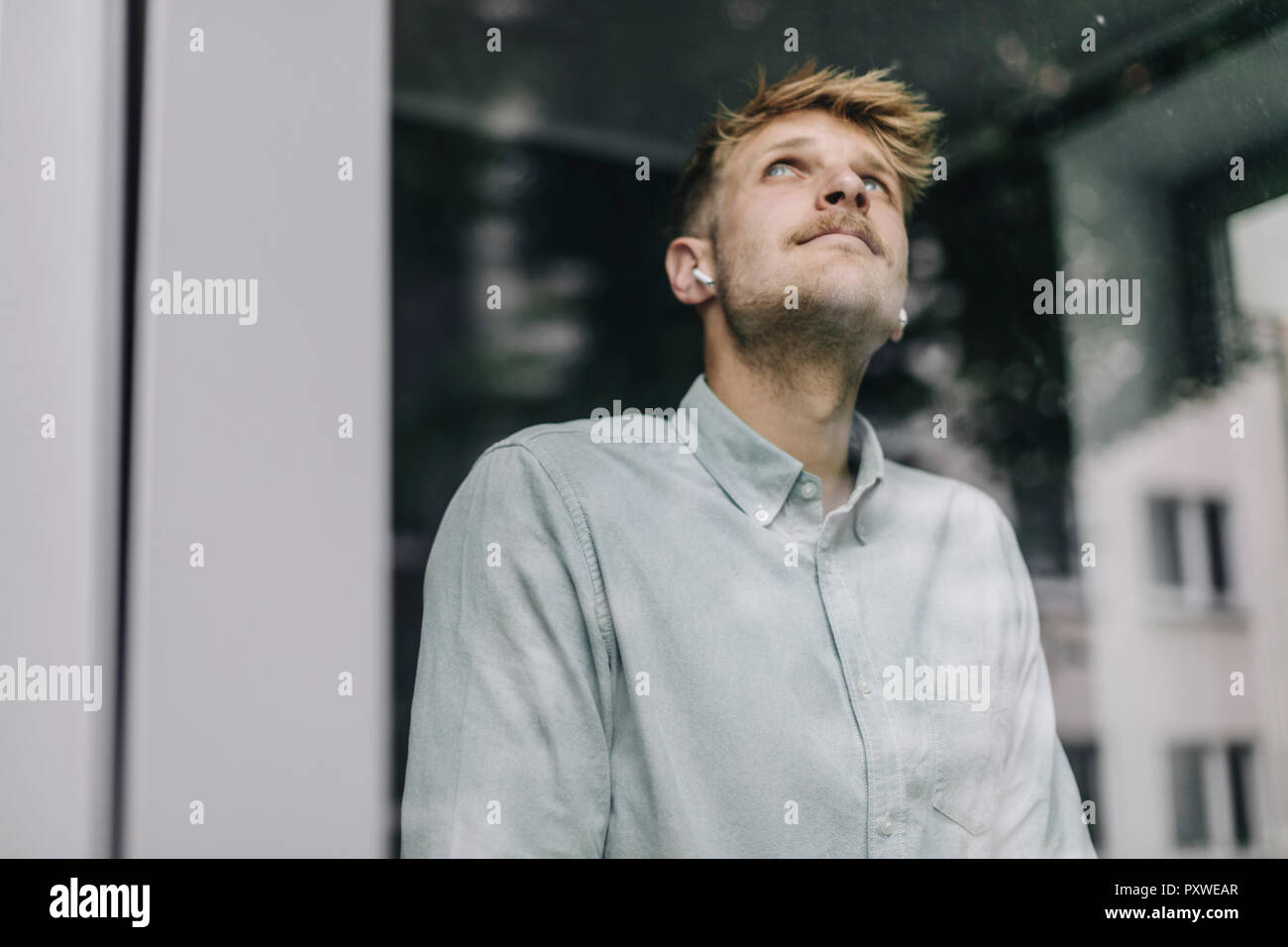 Jeune homme debout à la fenêtre, à l'aide d'oreillettes, daydreaming Banque D'Images