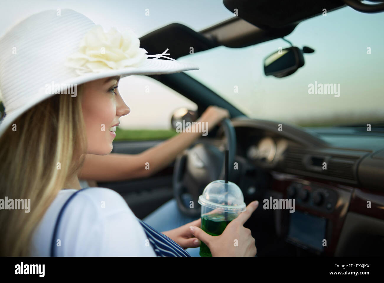 Vue de côté de la charmante jeune femme portant chapeau large bord blanc  avec fleur assis en cabriolet. Adorable Blonde modèle à la voiture et la  tenue de verre de limonade. Fille