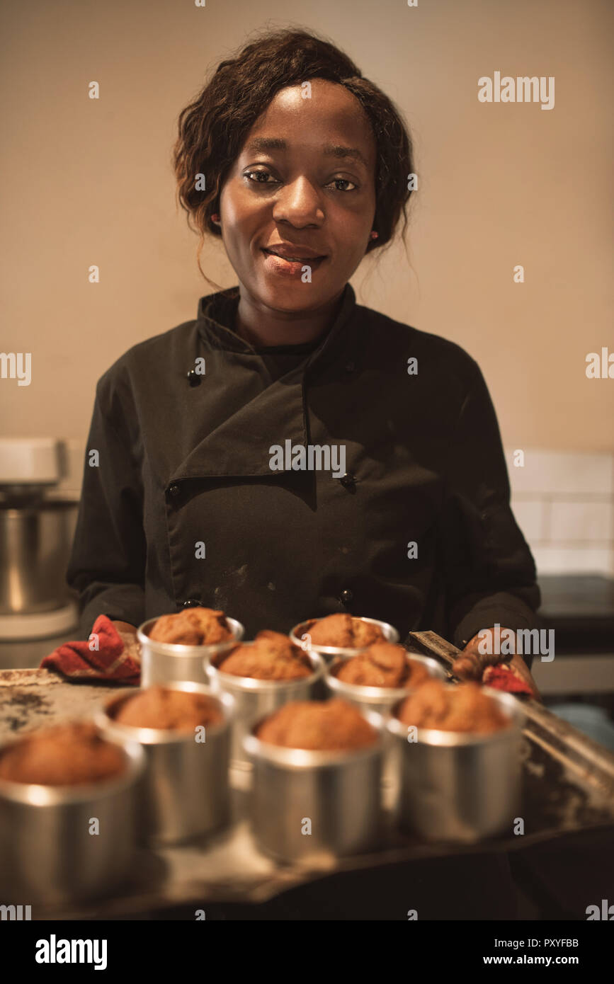 Smiling baker portant un plateau de muffins dans sa cuisine Banque D'Images