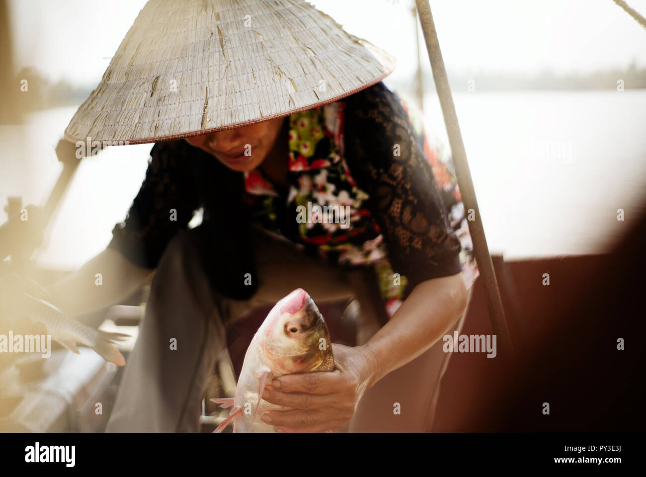 Pêcheuse vietnamiens en chapeau conique (non la) tenant un poisson et sortir du bateau quelque part sur le delta du fleuve Rouge Banque D'Images