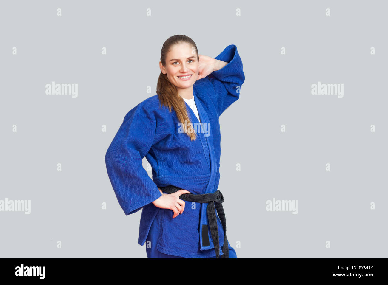 Beautiful happy karaté sport femme en bleu kimono avec ceinture noire holding main derrière sa tête sont à la recherche à l'appareil photo. Un martial japonais Banque D'Images