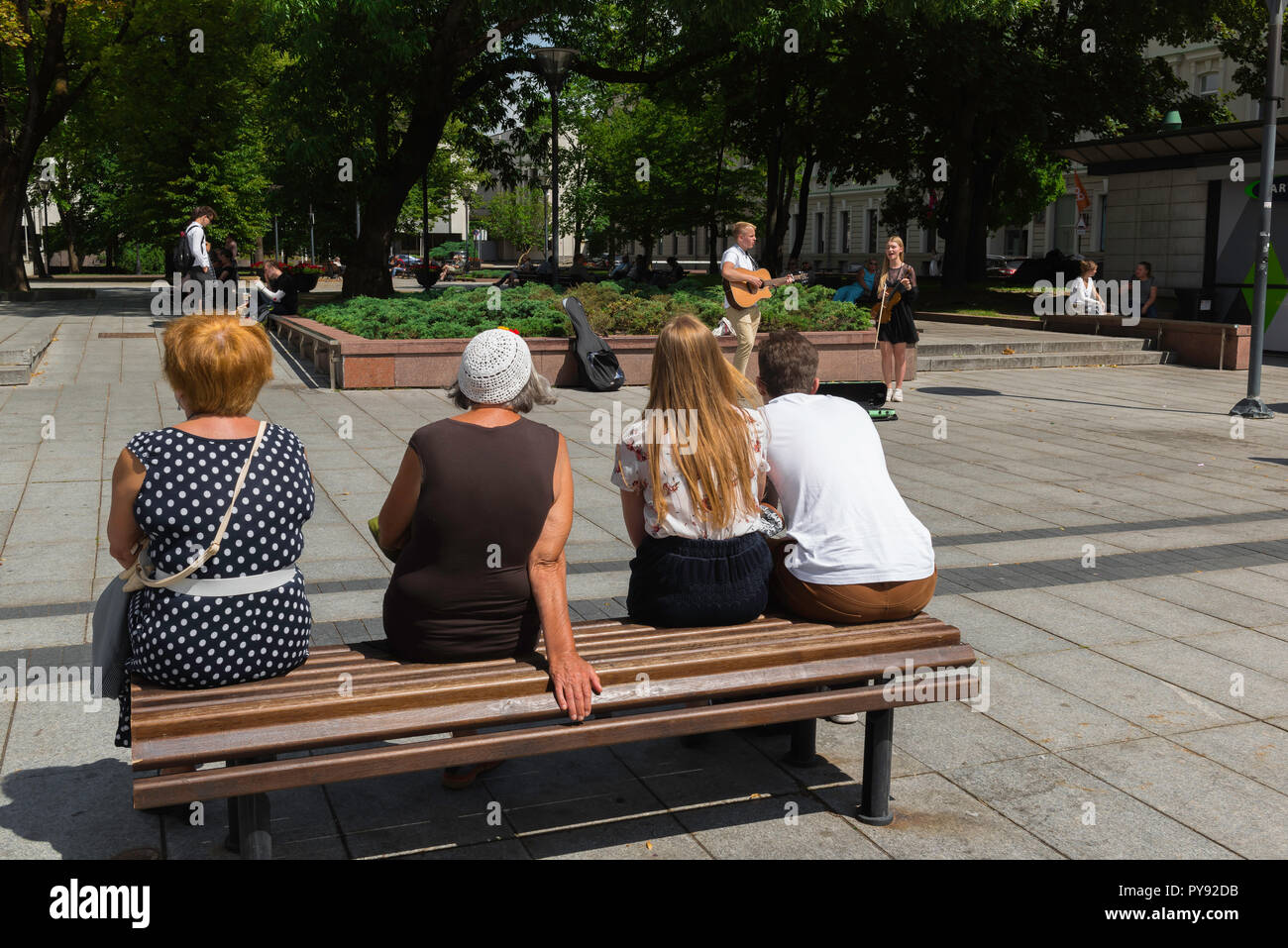 Vue arrière des gens assis sur un banc à regarder deux jeunes musiciens dans la rue Gedimino Prospektas à Vilnius, Lituanie. Banque D'Images