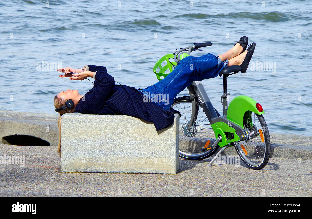 Jeune femme aux cheveux longs, broche-blouson rayé et d'un blue-jean, allongé sur un mur de pierre, à côté de la Seine aux pieds reposant sur un vélo loué velib' Banque D'Images