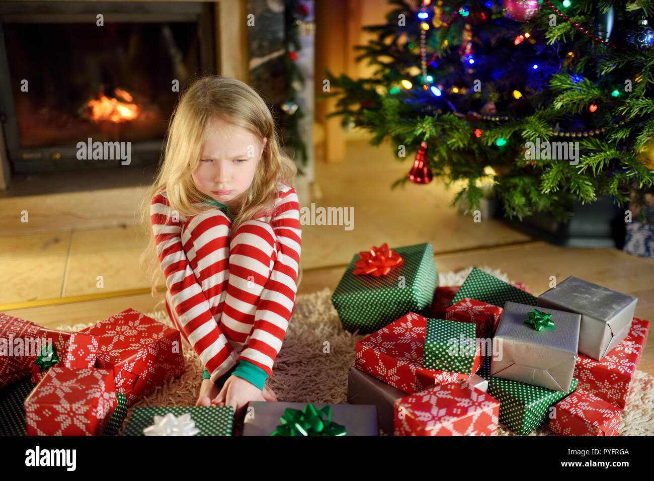 Cute little girl se sentir malheureux avec ses cadeaux de Noël. Enfant  assis par une cheminée dans un confortable salon sombre sur veille de Noël. Trop  de cadeaux pour Photo Stock -