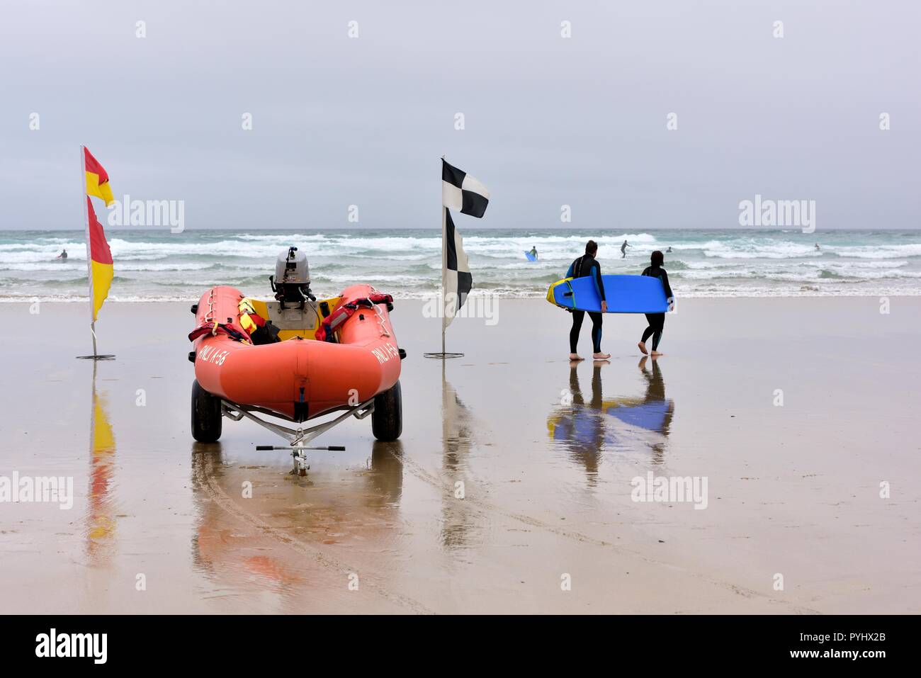 Les personnes portant un blue surf board en passant devant la plage de la RNLI embarcation gonflable et drapeaux de sécurité,Broad Oak Beach, Cornwall, Angleterre, Royaume-Uni Banque D'Images