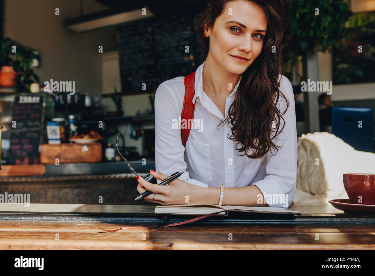 Belle femme assise à un café en regardant la caméra. Certain modèle féminin portant une chemise blanche détente au café avec son journal sur la table. Banque D'Images